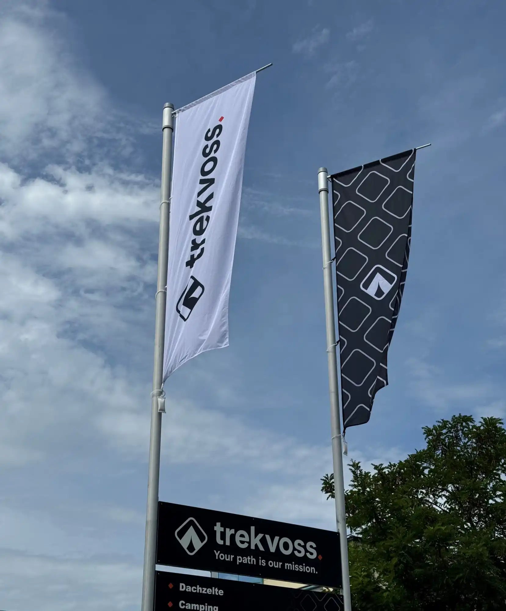 Zwei hohe Werbefahnen, die die Marke und das Logo von Trekvoss vor einem blauen Himmel zeigen.