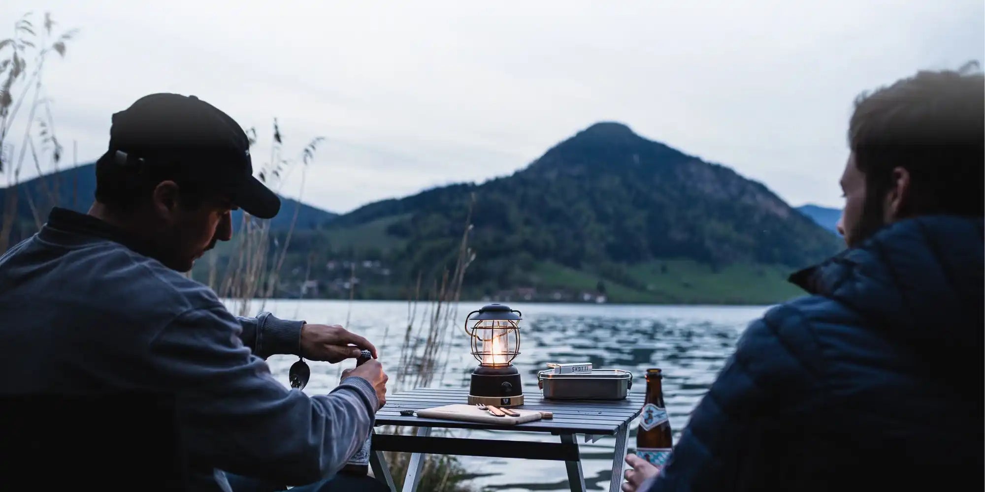 Zwei Personen sitzen an einem Tisch mit einer Laterne, die auf einen See und einen Berg blickt.