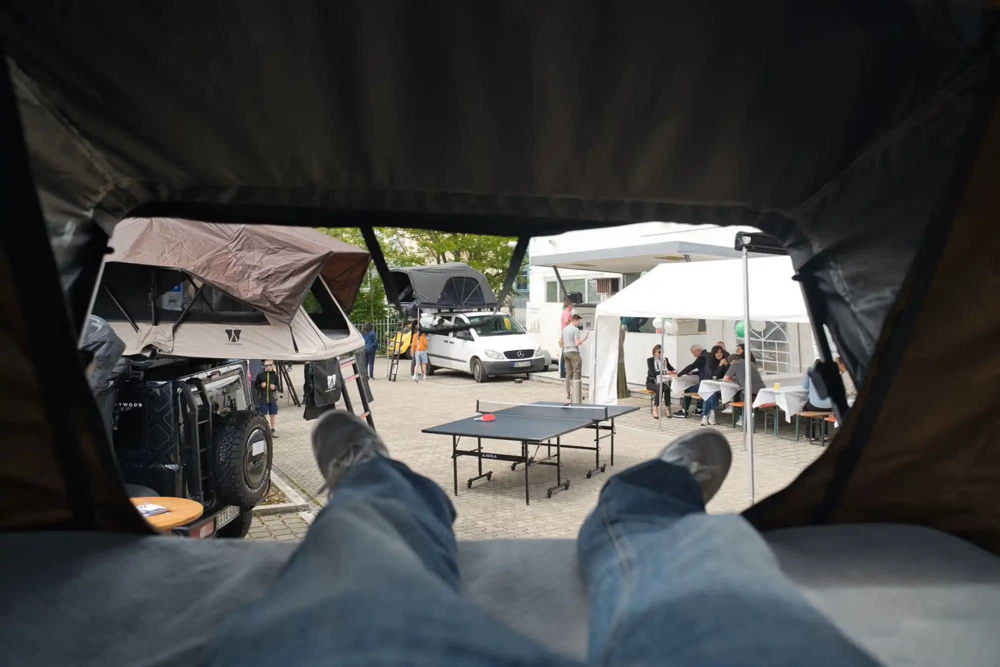 Die Beine und Füße einer Person, die in einem Dachzelt ruhen, während sie bei einer Outdoor-Versammlung mit Fahrzeugen und einem Tischtennistisch zuschaut.